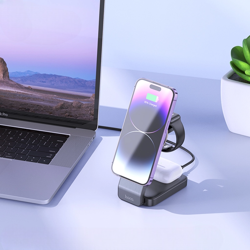 Wireless charging stand with smartphone and earbuds on a desk next to a laptop